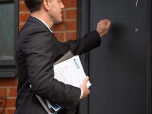 Smartly dressed man knocking at front door with DCBL papers